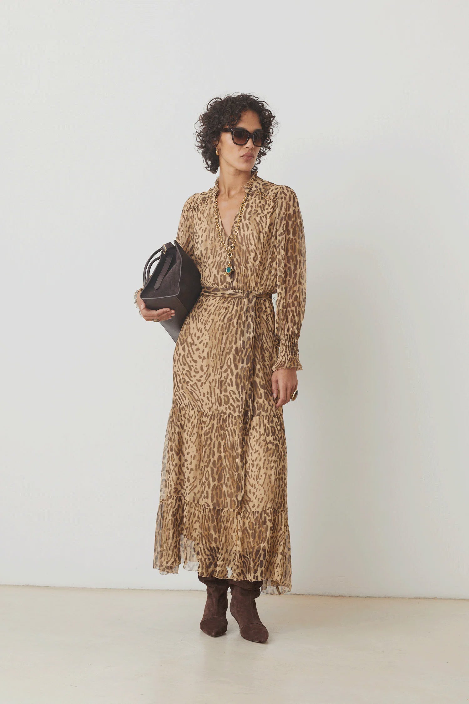 A woman with short curly hair, sunglasses, and brown boots models the SUNCOO Cosmos Dress—a loose-fit leopard print viscose dress—while holding a black handbag against a plain white background.