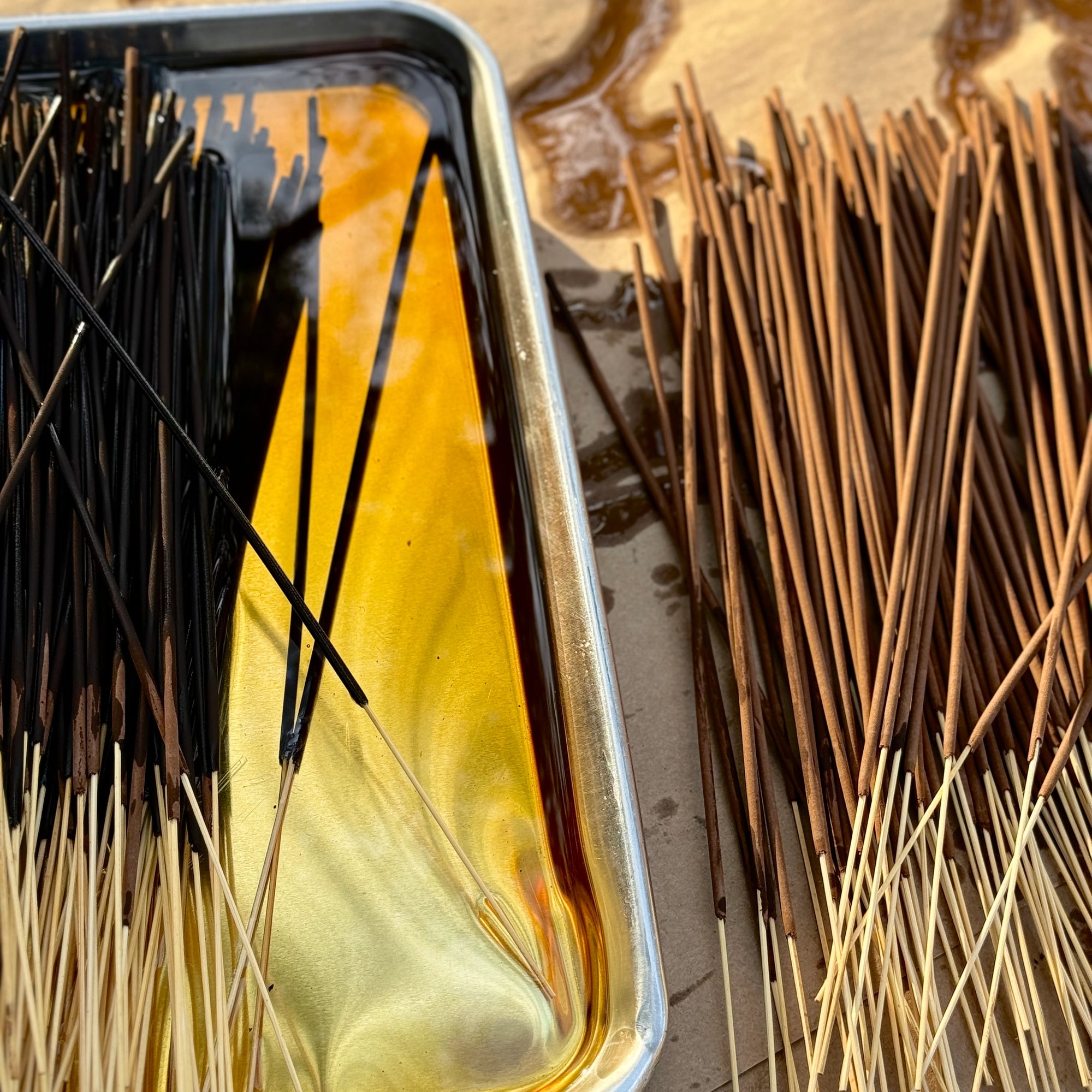 Close-up of Faire Incense Sticks being dipped in yellow sandalwood fragrance oil on a silver tray, with wet and dry sticks neatly arranged side by side.