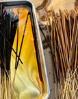 Close-up of Faire Incense Sticks being dipped in yellow sandalwood fragrance oil on a silver tray, with wet and dry sticks neatly arranged side by side.