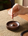 A hand places a lit incense stick into the Faire Walnut Wood Incense Burner on a woven placemat, with smoke rising gently—showcasing traditional Chinese artistry and a unique rustic glaze.