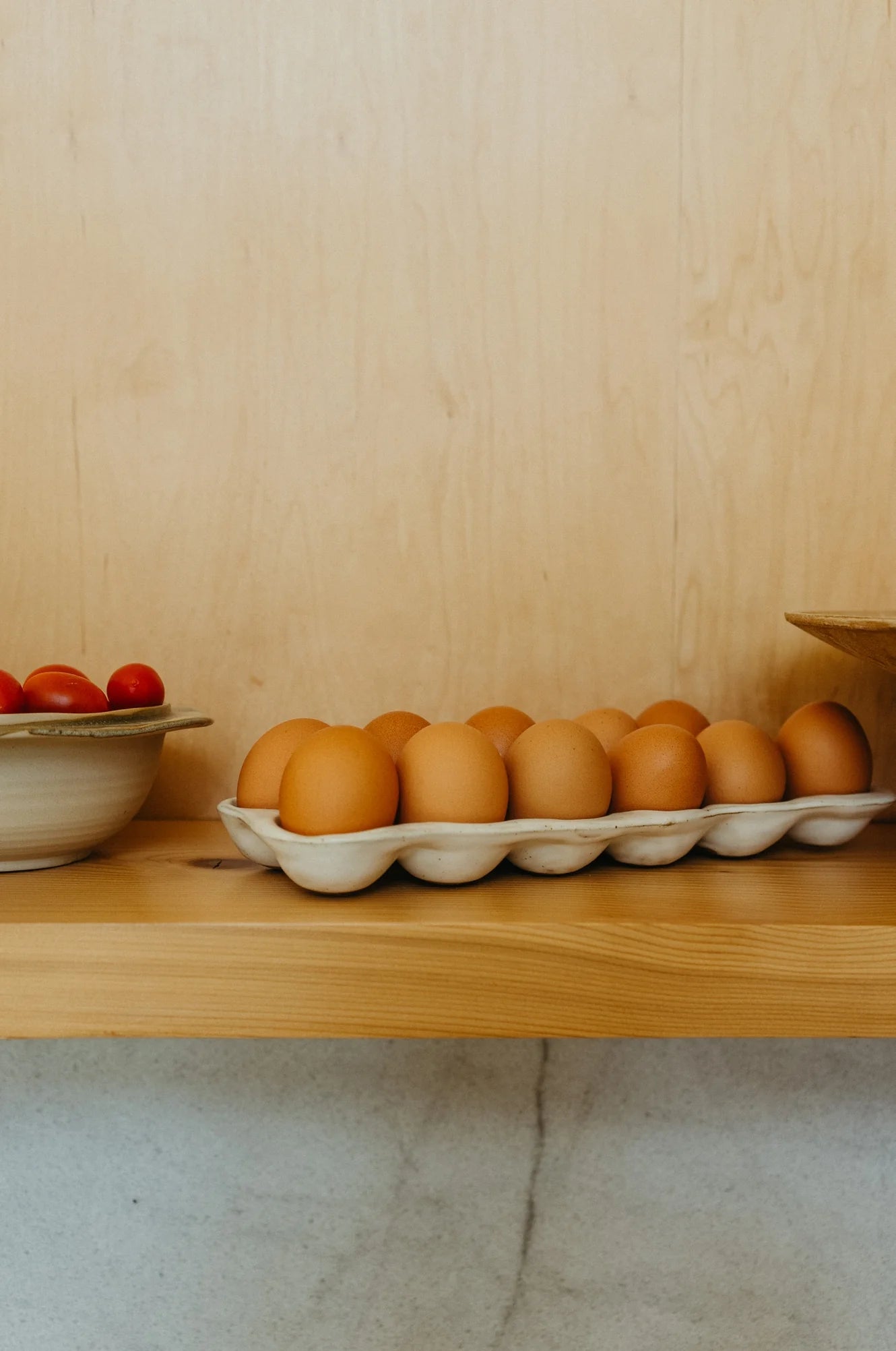 The Indaba Homestead Egg Holder displays brown eggs on a wooden shelf, serving as stylish storage and decor, placed beside a bowl of red cherry tomatoes against a light wood background.