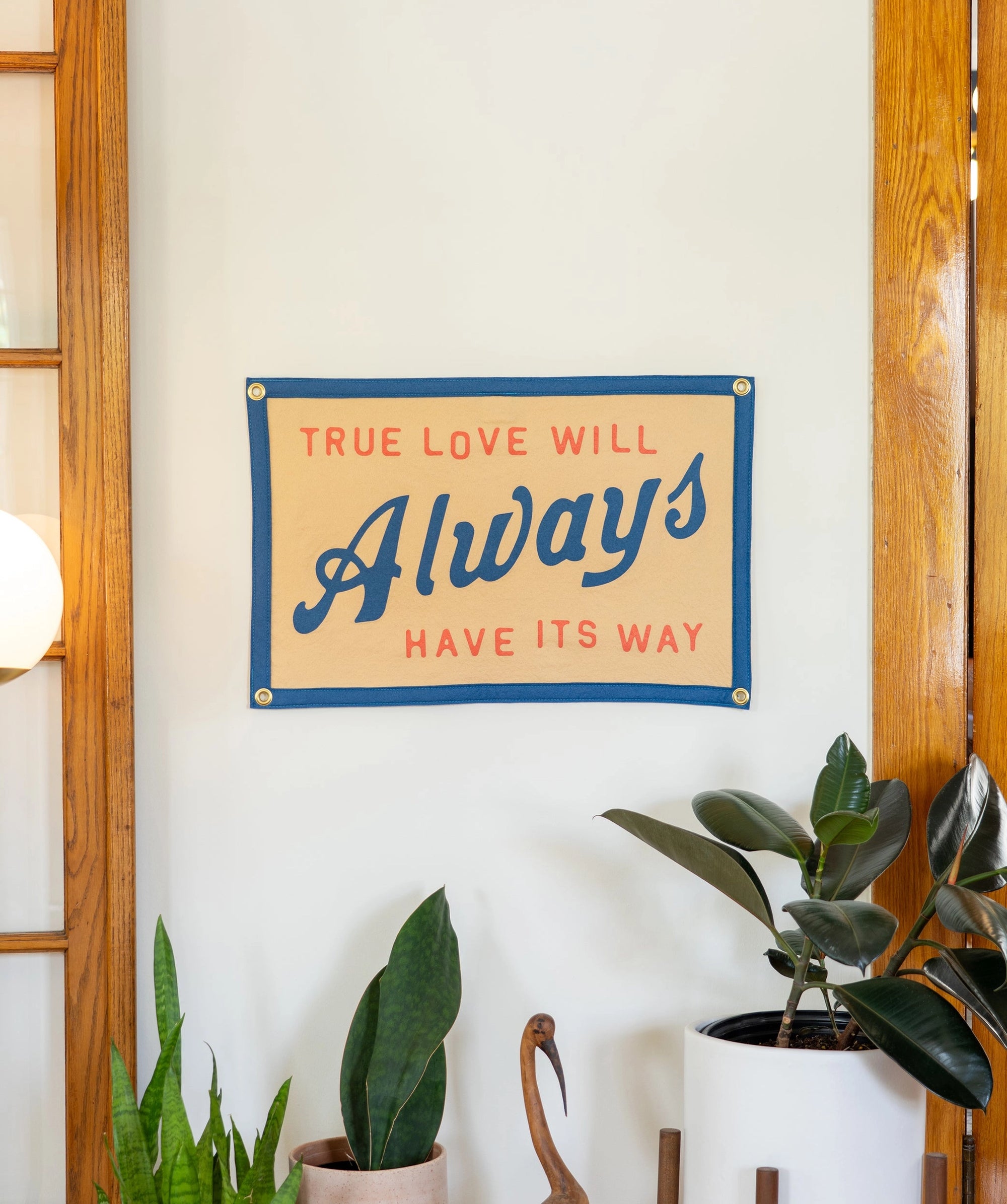 The Oxford Pennant "True Love Will Always Have It's Way" felt wall hanging features red and blue text, inspired by John Prine’s lyrics. Displayed above green plants and near wood-framed glass doors, it brightens any white wall.