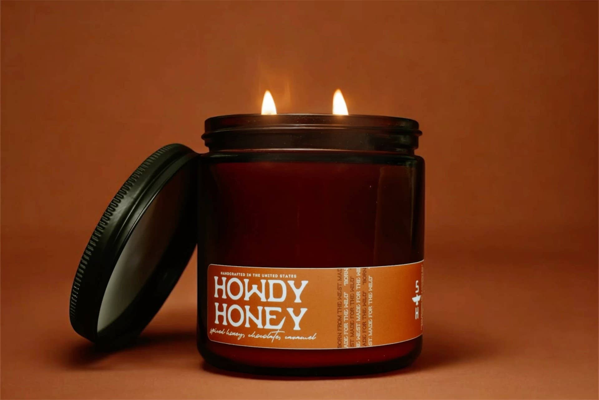 The Faire Howdy Honey Candle is a brown coconut soy wax jar candle with two flames, shown beside its lid on a warm brown background.