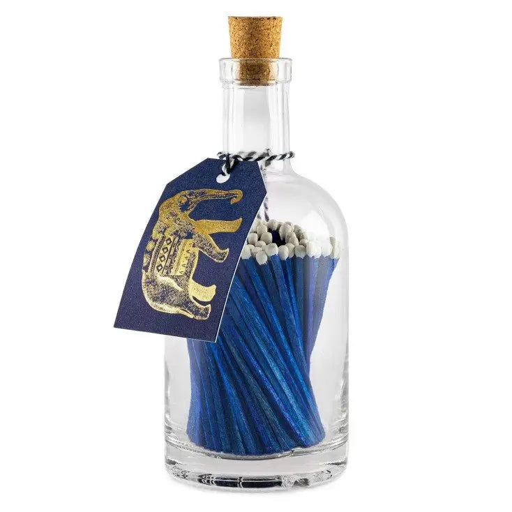 The Faire Glass Bottle Matches offer sustainable home living with blue matches and white tips in a cork-sealed glass bottle. A blue tag featuring a gold elephant is attached to the neck with black and white string, all in plastic-free packaging.