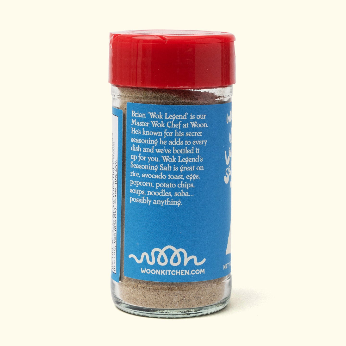 A glass jar with a red lid contains Faire’s Woon Wok Legend's Seasoning Salt. The blue label suggests using it on rice, eggs, popcorn, chips, and soup, displays "woonkitchen.com," and shares a brief Master Wok Chef bio.