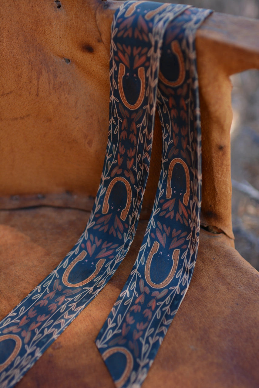 The Faire Western Harvest Twilly scarf features hand-drawn horseshoe and leafy vine patterns in brown and gold tones, displayed over a rustic brown leather chair.