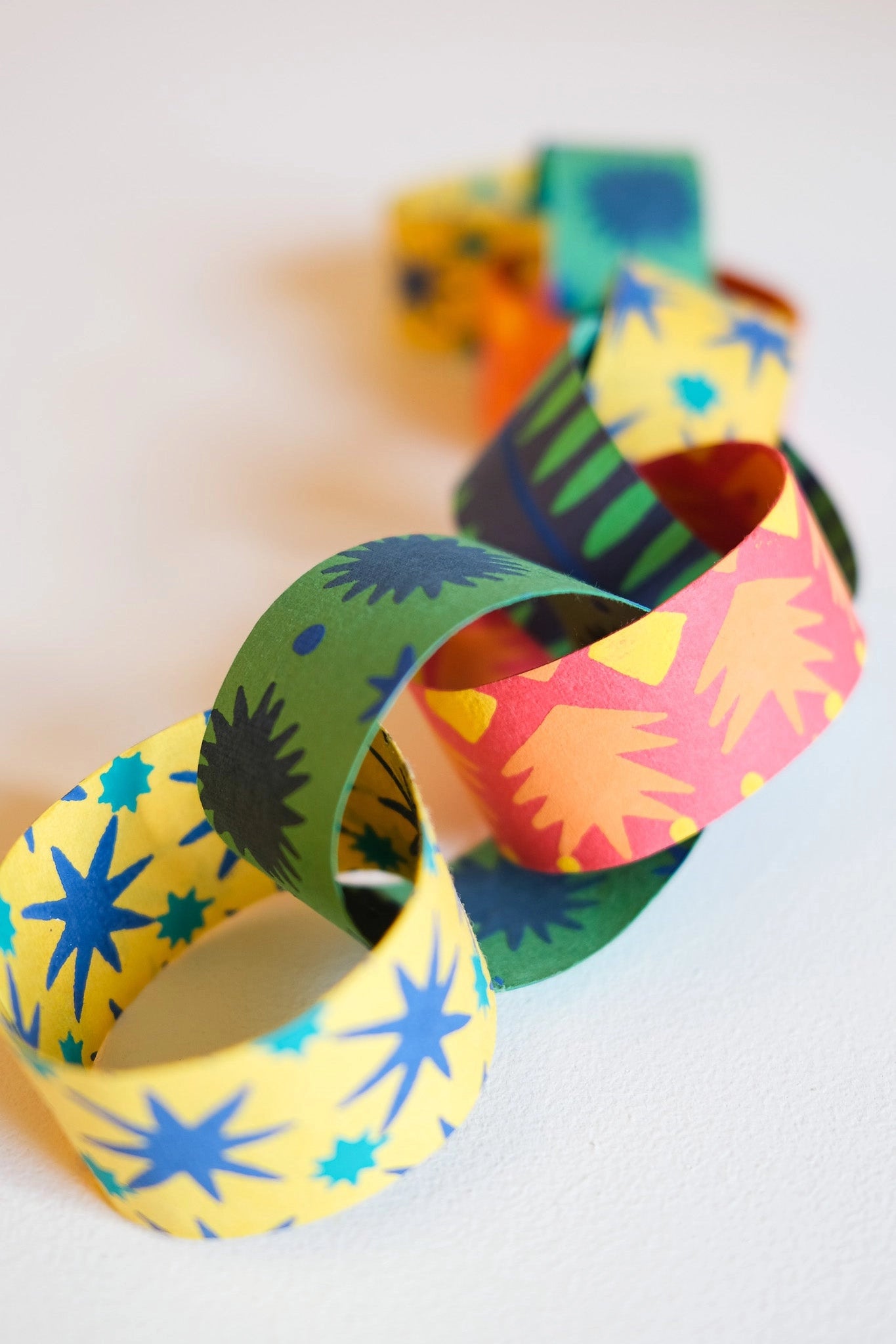 The Faire Paper Garland/Chains feature interlocking loops with vibrant starburst and sunburst patterns in yellow, green, blue, and red—perfect for festive birthday decorations or garlands, beautifully displayed on a white surface.