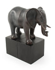 The Zentique Elephant Bookends feature a dark bronze elephant standing with one foot on each of two black rectangular blocks, making them a unique and stylish decorative bookend set.