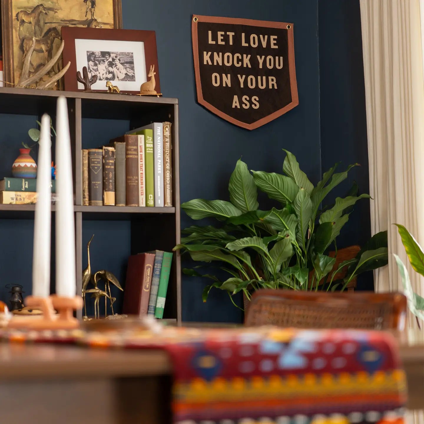 A cozy room features a bookshelf, potted plant, and colorful tablecloth. On the dark blue wall hangs Faire’s "Let Love Knock You" Camp Flag, boldly declaring some classic relationship advice.