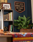 A cozy room features a bookshelf, potted plant, and colorful tablecloth. On the dark blue wall hangs Faire’s "Let Love Knock You" Camp Flag, boldly declaring some classic relationship advice.