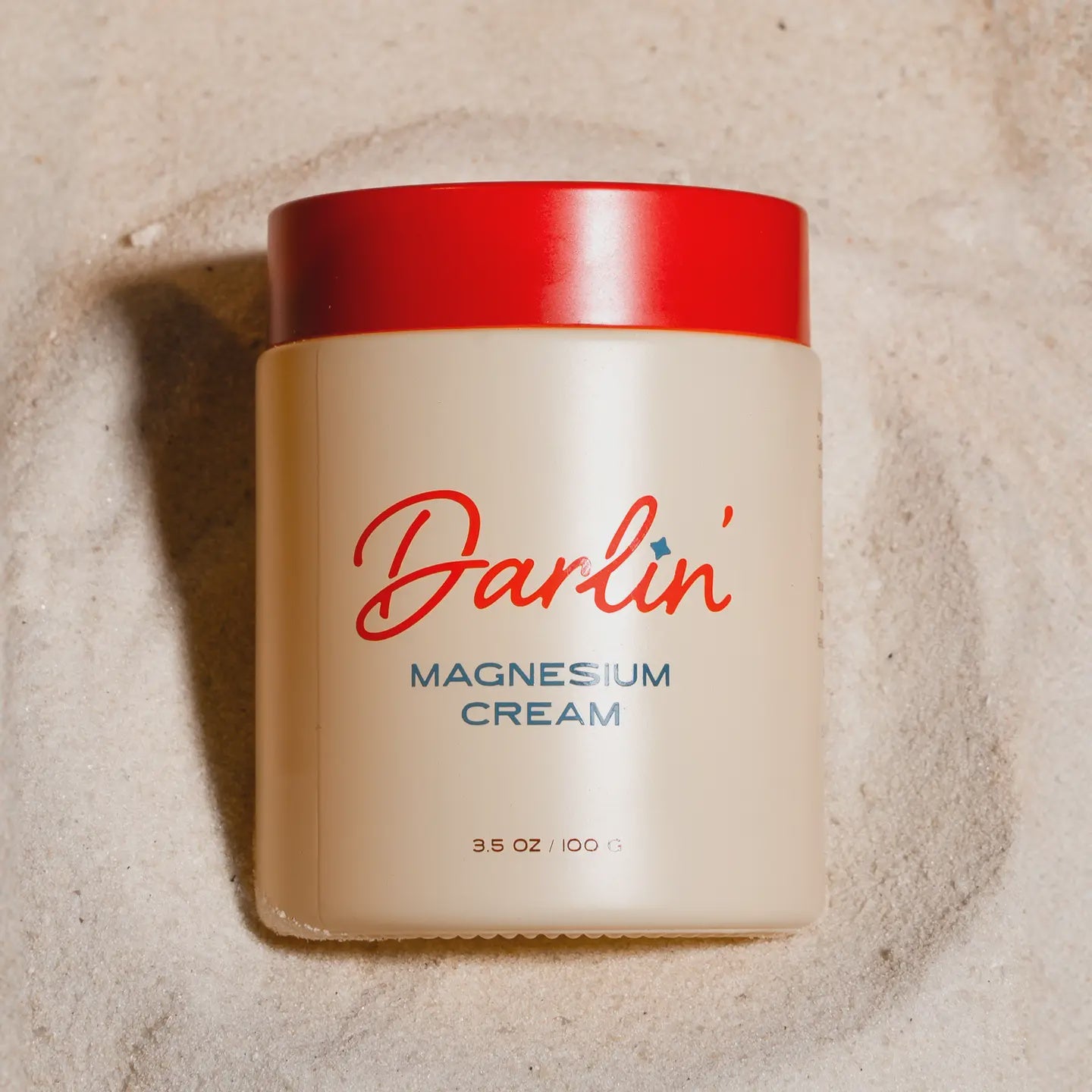 A jar of Faire Magnesium Cream Tallow with a red lid rests on sand. Its cream-colored label, featuring red and blue text, showcases 3.5 ounces (100 grams) of natural skincare, ideal for those prioritizing tallow in their routine.