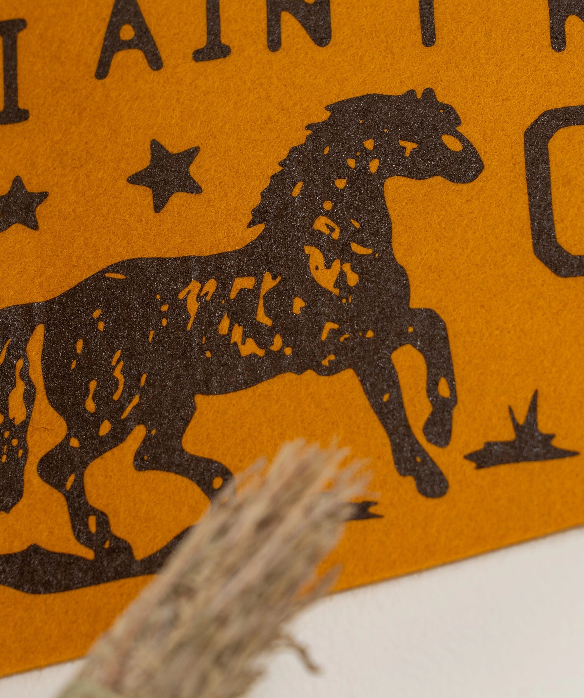 Close-up of Faire's "I Ain't No Cowboy Pennant" in brown felt, with an old gold hand-drawn horse galloping among stars and partial text; a bundle of dried grass appears in the foreground.