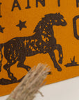 Close-up of Faire's "I Ain't No Cowboy Pennant" in brown felt, with an old gold hand-drawn horse galloping among stars and partial text; a bundle of dried grass appears in the foreground.