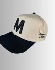 The M Hat by Faire features beige and black embroidery, a bold black "M" on the front, and "Strong as a Mother" stitched on the side, shown against a light gray gradient background.