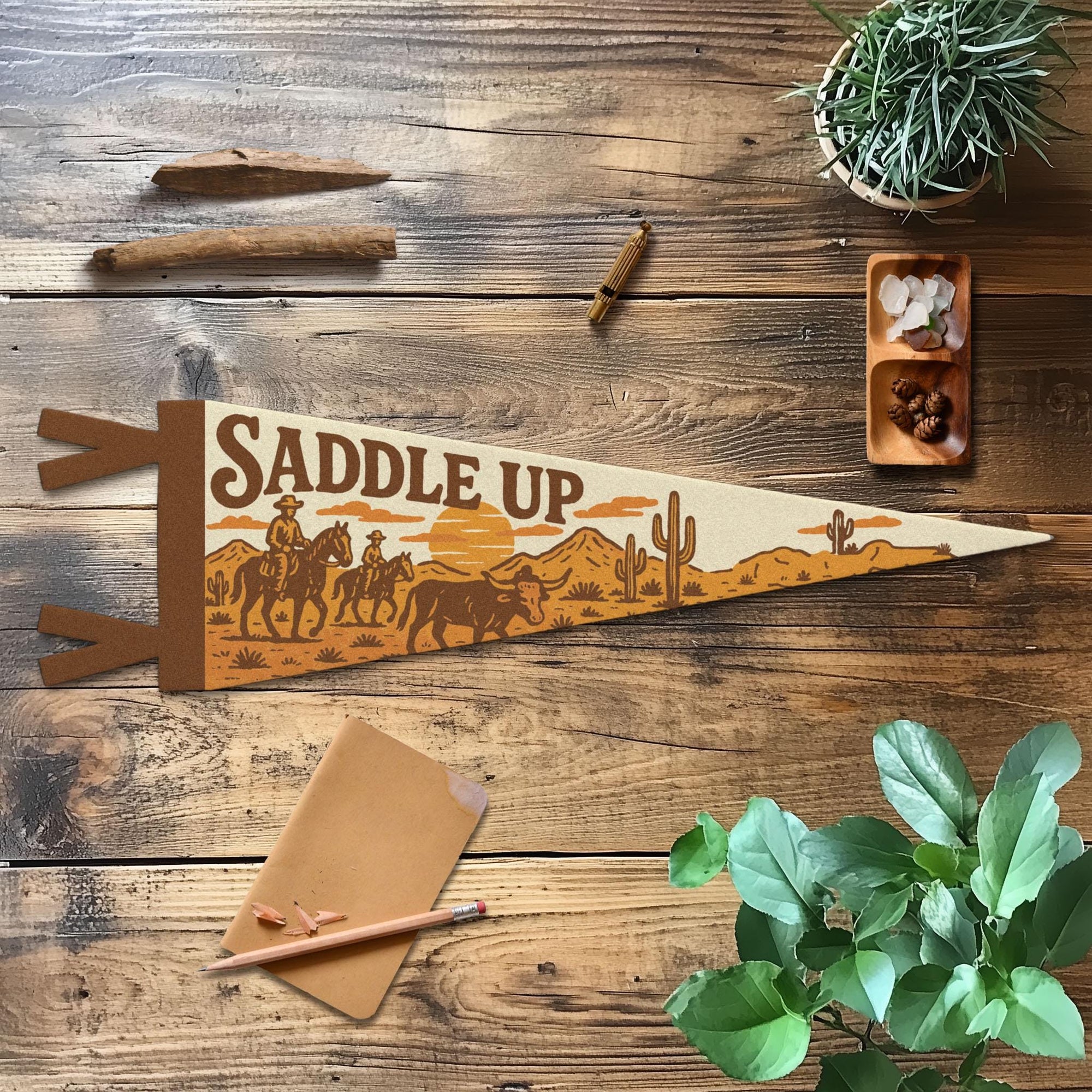 The "Saddle Up" Felt Pennant by Faire features a cowboy scene and lies on a wooden table with plants, pencil, cinnamon stick, crystals, and a notepad—perfect for western decor or as a unique gift for horse lovers.