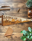The "Saddle Up" Felt Pennant by Faire features a cowboy scene and lies on a wooden table with plants, pencil, cinnamon stick, crystals, and a notepad—perfect for western decor or as a unique gift for horse lovers.