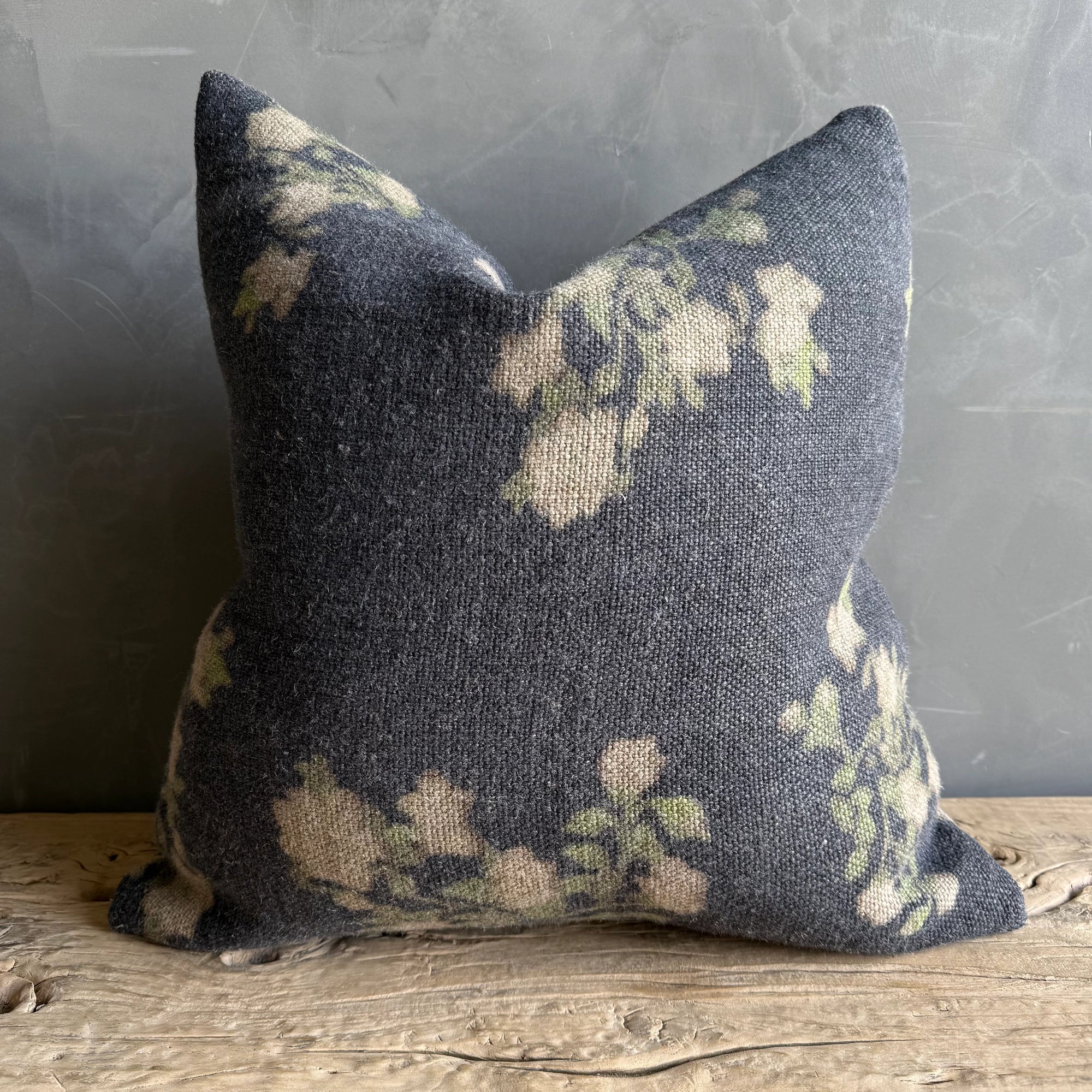 The Faire French Linen Roses Pillow, 26x26, features dark gray fabric with a soft faded floral pattern in light cream and green. Placed on a rustic wooden surface against a textured gray wall, it brings charming floral accents to any space.