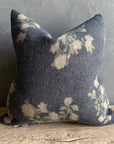 The Faire French Linen Roses Pillow, 26x26, features dark gray fabric with a soft faded floral pattern in light cream and green. Placed on a rustic wooden surface against a textured gray wall, it brings charming floral accents to any space.