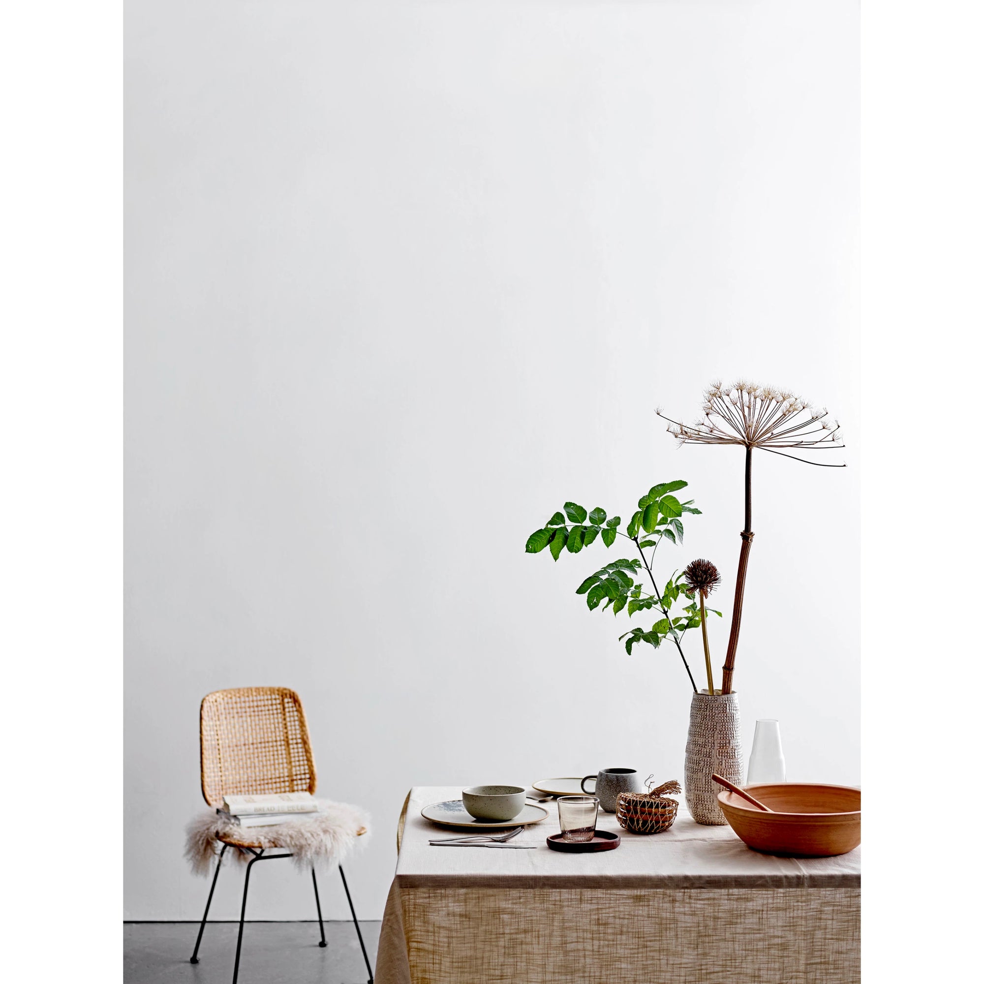 A minimalist dining scene in a Scottsdale, Arizona bungalow, featuring a wooden table covered by a beige tablecloth, adorned with various bowls and a cup. A woven chair with a white fluffy throw sits beside it, next to a Bloomingville stoneware vase with tall dried flowers.