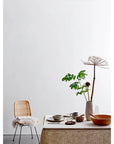 A minimalist dining scene in a Scottsdale, Arizona bungalow, featuring a wooden table covered by a beige tablecloth, adorned with various bowls and a cup. A woven chair with a white fluffy throw sits beside it, next to a Bloomingville stoneware vase with tall dried flowers.