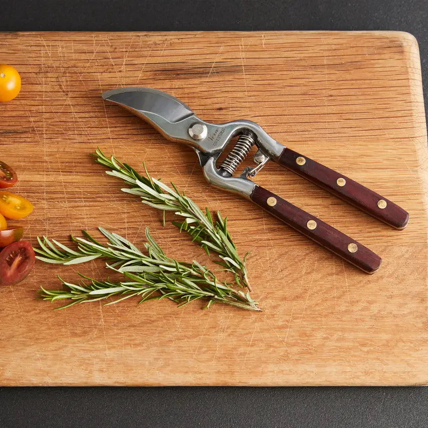 The Faire Thai Kitchen Garden Shears, featuring stainless steel blades and wooden handles, rest on a cutting board alongside fresh rosemary sprigs and sliced cherry tomatoes.
