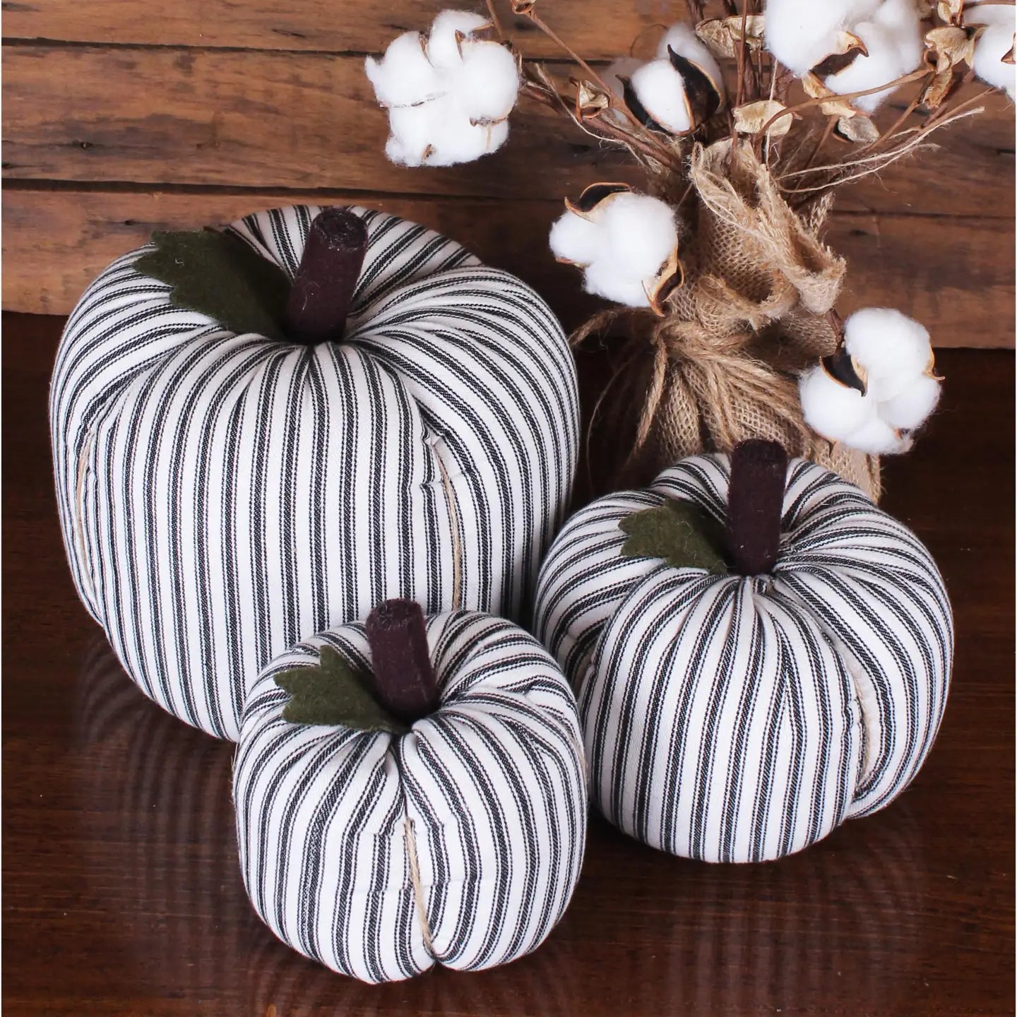 The Faire Pumpkin set features three black-and-white striped fabric pumpkins on a wooden surface beside burlap-wrapped cotton stems, creating charming farmhouse decor—perfect autumn ornaments.