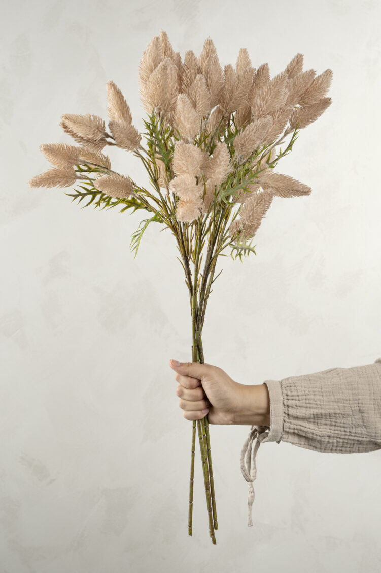 A hand holds the Indaba Pussytail Grass Spray, a bouquet of beige long-stem faux plants, against a light textured background. The person wears a light-colored, long-sleeved shirt with tied cuffs.