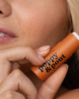 A person holds a Poppy & Pout Lip Balm by Faire near their lips, emphasizing the eco-friendly product's label and focusing on their lips.