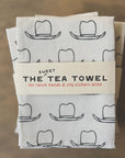 A folded white tea towel featuring black cowboy hats is wrapped in a cotton belly band. The label states "THE SWEET TEA TOWEL by Faire, for ranch hands & city slickers alike," emphasizing its ethically-made quality.