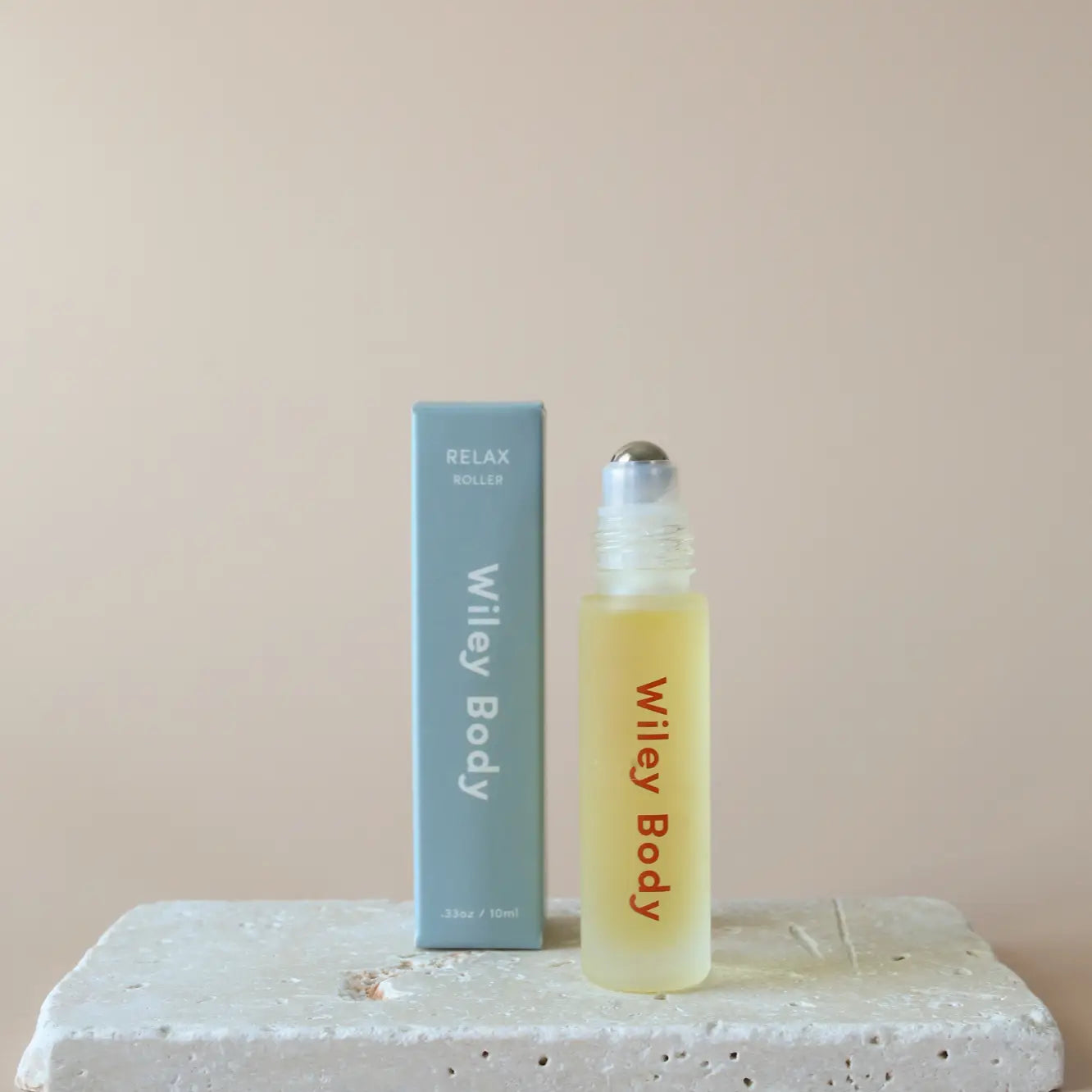 A small Wiley Roller with a metal top rests beside its light blue box on a textured stone, set against a beige background, embodying well-being through calming essential oils.