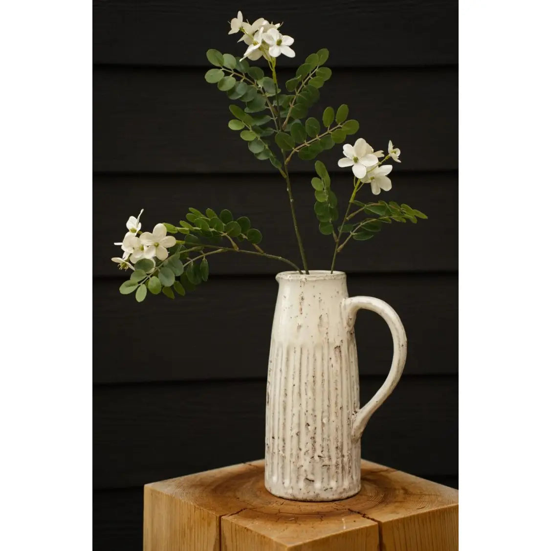 The Indaba Locust Flower Branch features green stems arranged in a rustic white ceramic pitcher with vertical grooves, set on a wooden block against a dark paneled backdrop—an ideal accent decor for any space.
