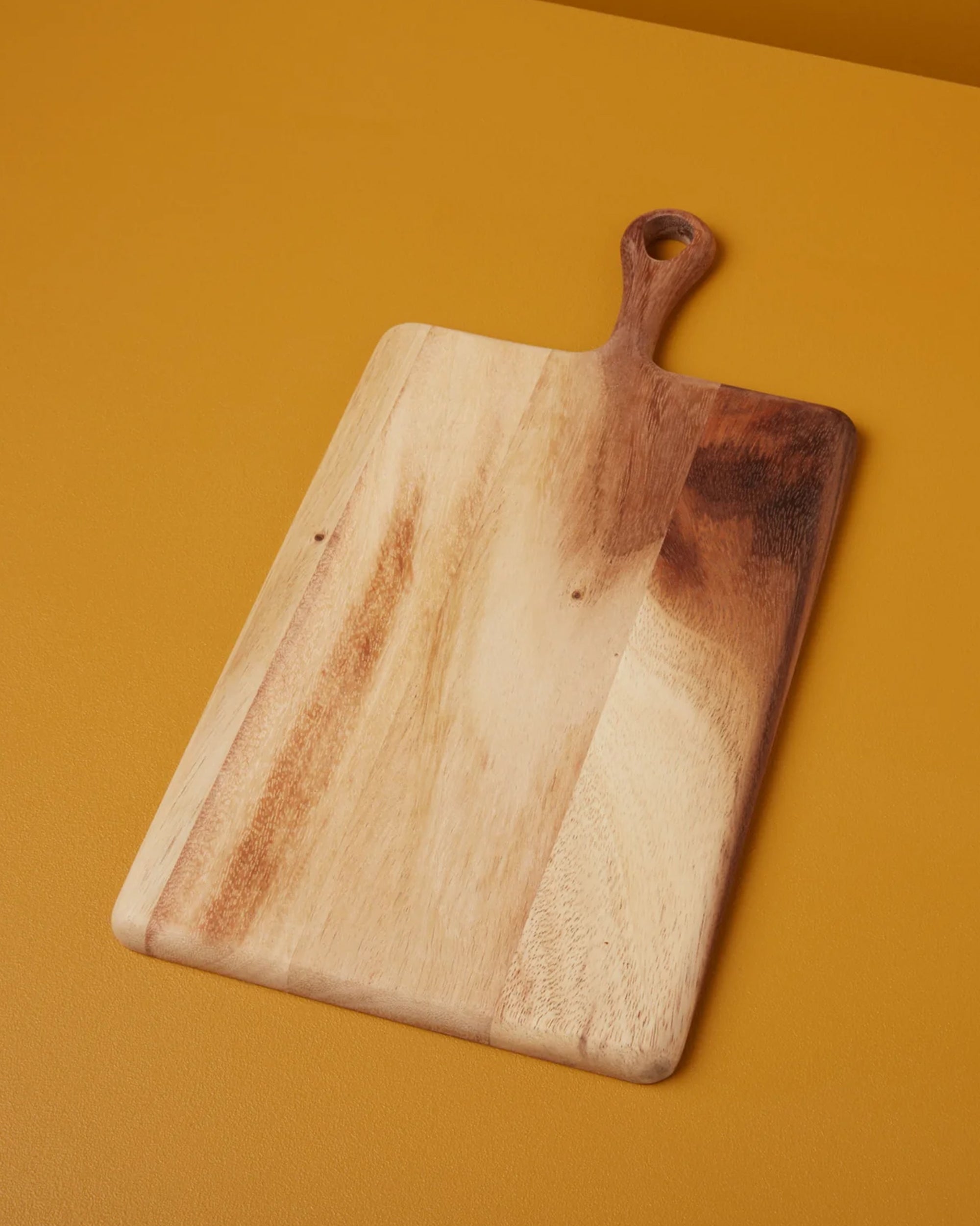 The Be Home Acacia Wood Rec Board S, a handcrafted rectangular board with handle and hanging hole, displays natural wood grain patterns as it rests on a mustard yellow surface.