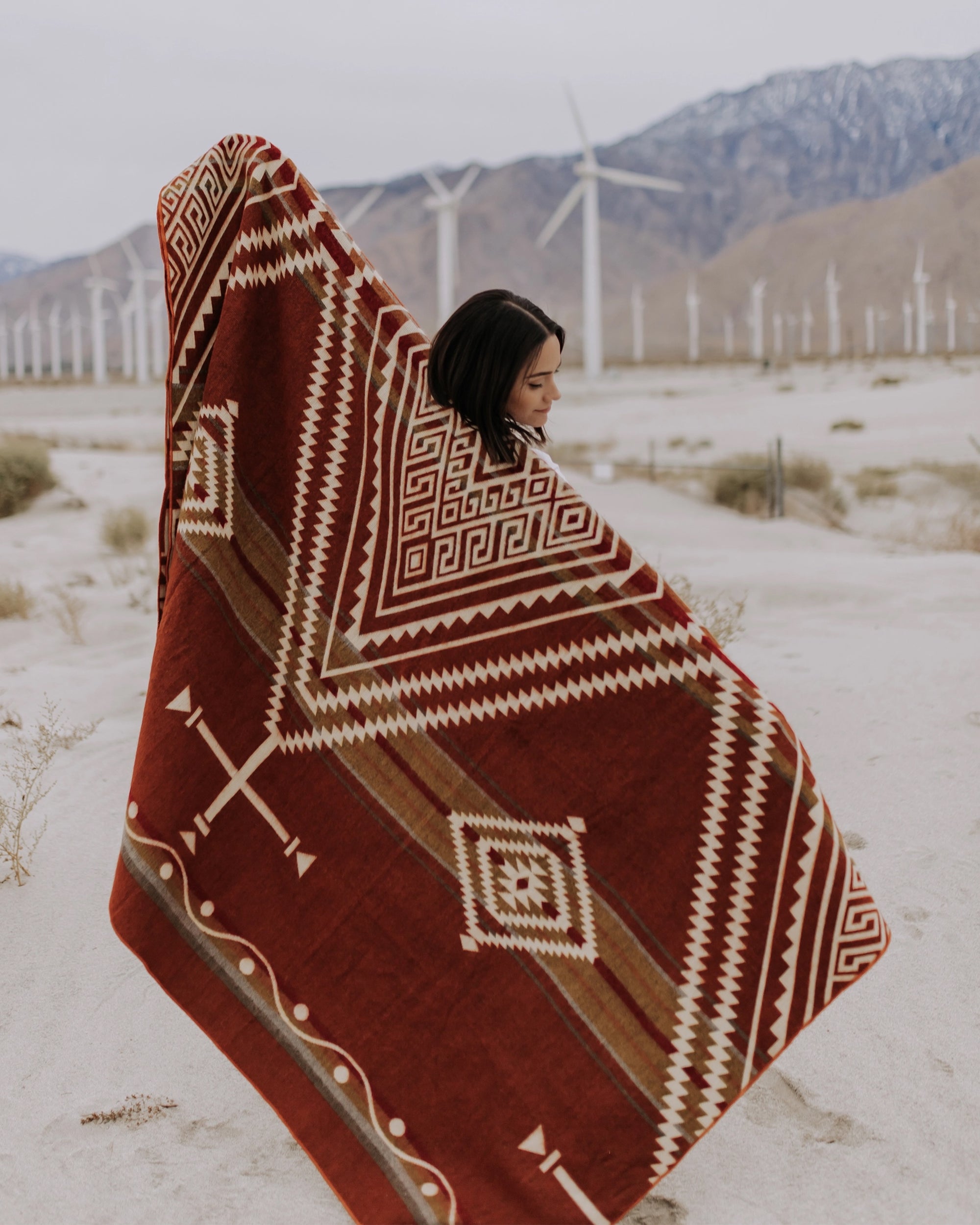 A person stands in a sandy landscape wrapped in the Achuar Blanket by Faire, with wind turbines and mountains in the background, creating a serene scene with boho decor vibes.