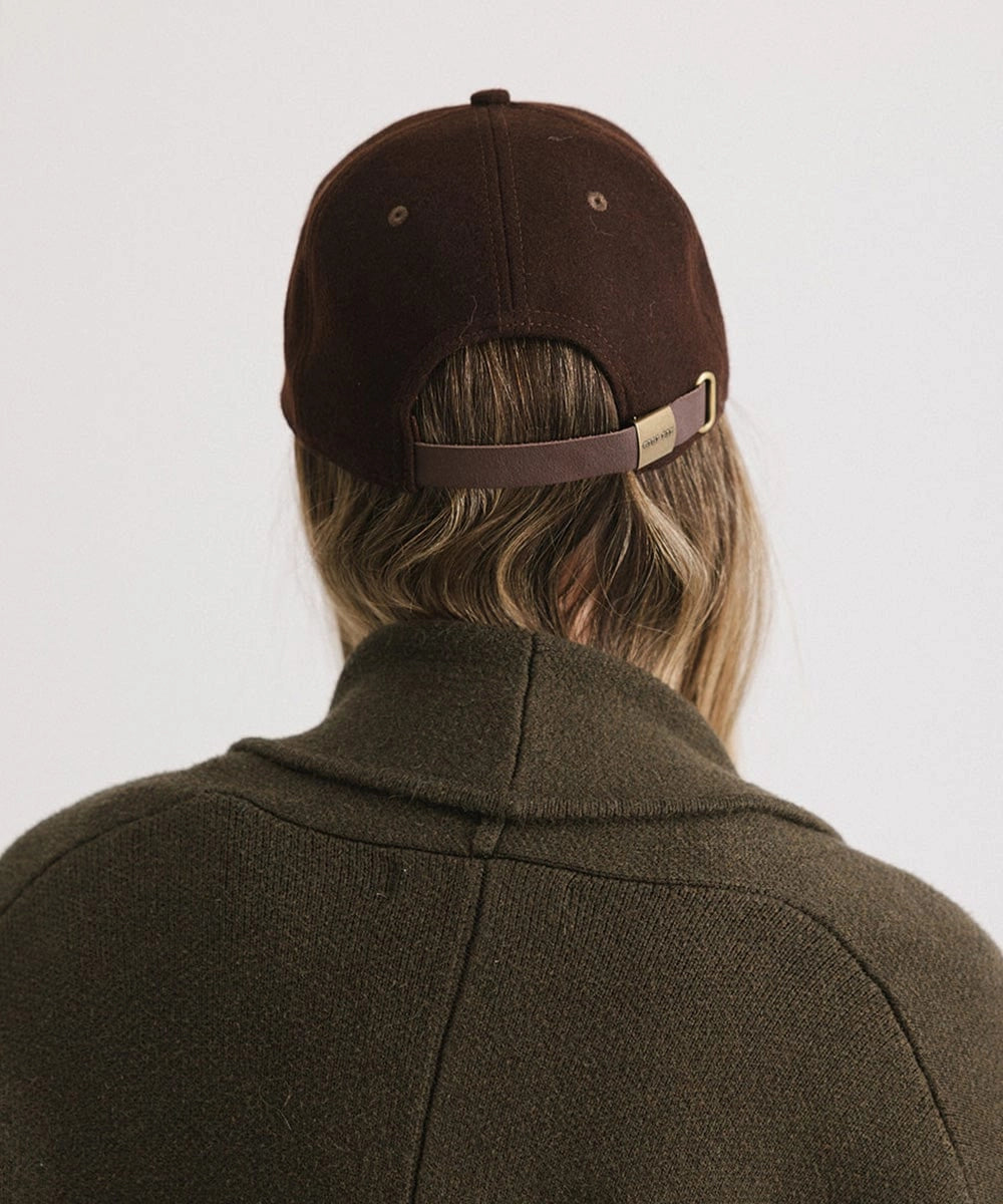 Someone with long hair is seen from behind wearing the Faire Aven Leather Ball Cap in dark brown, paired with an olive green sweater, set against a plain light background—a chic winter accessory.
