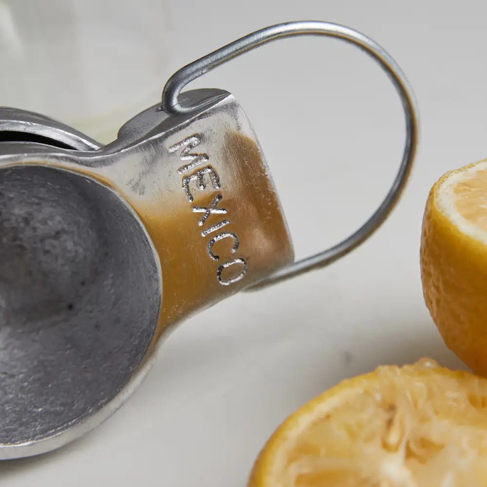 Close-up of a Faire Aluminum Hand Juicer embossed with &quot;MEXICO,&quot; surrounded by halved lemons and limes on a neutral surface.