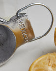 Close-up of a Faire Aluminum Hand Juicer embossed with "MEXICO," surrounded by halved lemons and limes on a neutral surface.