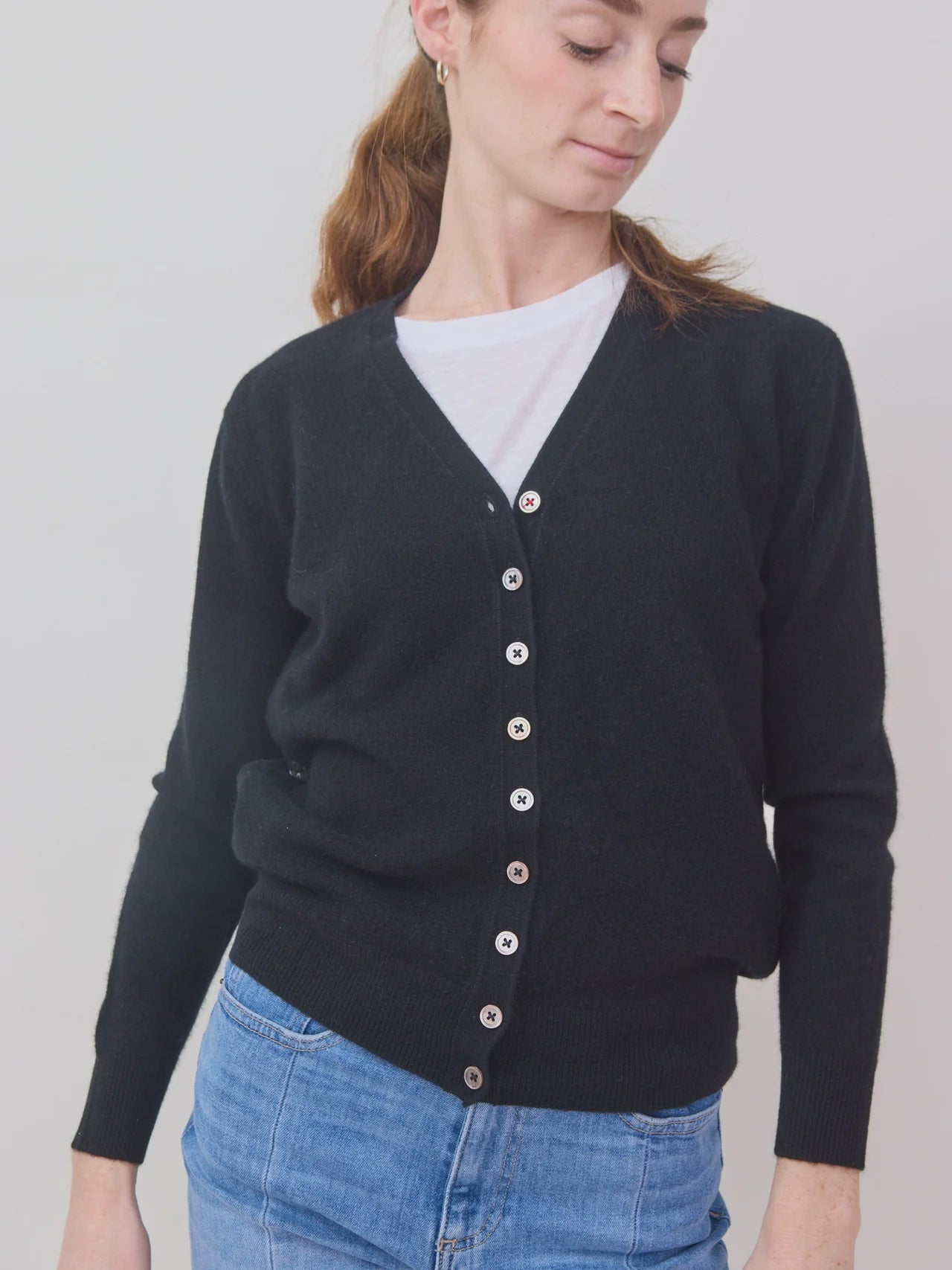 A woman wears the Brazeau Tricot Tulle Back Cardigan in black over a white T-shirt and blue jeans, standing against a plain light background and looking down to the side with a slight smile.