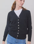 A woman wears the Brazeau Tricot Tulle Back Cardigan in black over a white T-shirt and blue jeans, standing against a plain light background and looking down to the side with a slight smile.