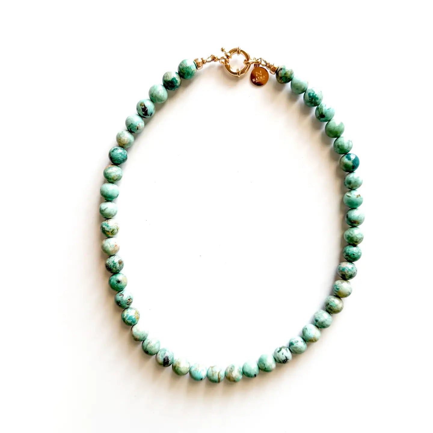 The Faire Galleria Necklace is a 17-inch gold-plated piece with round, light turquoise-green beaded stones and a gold clasp, displayed in a circle on a white background.