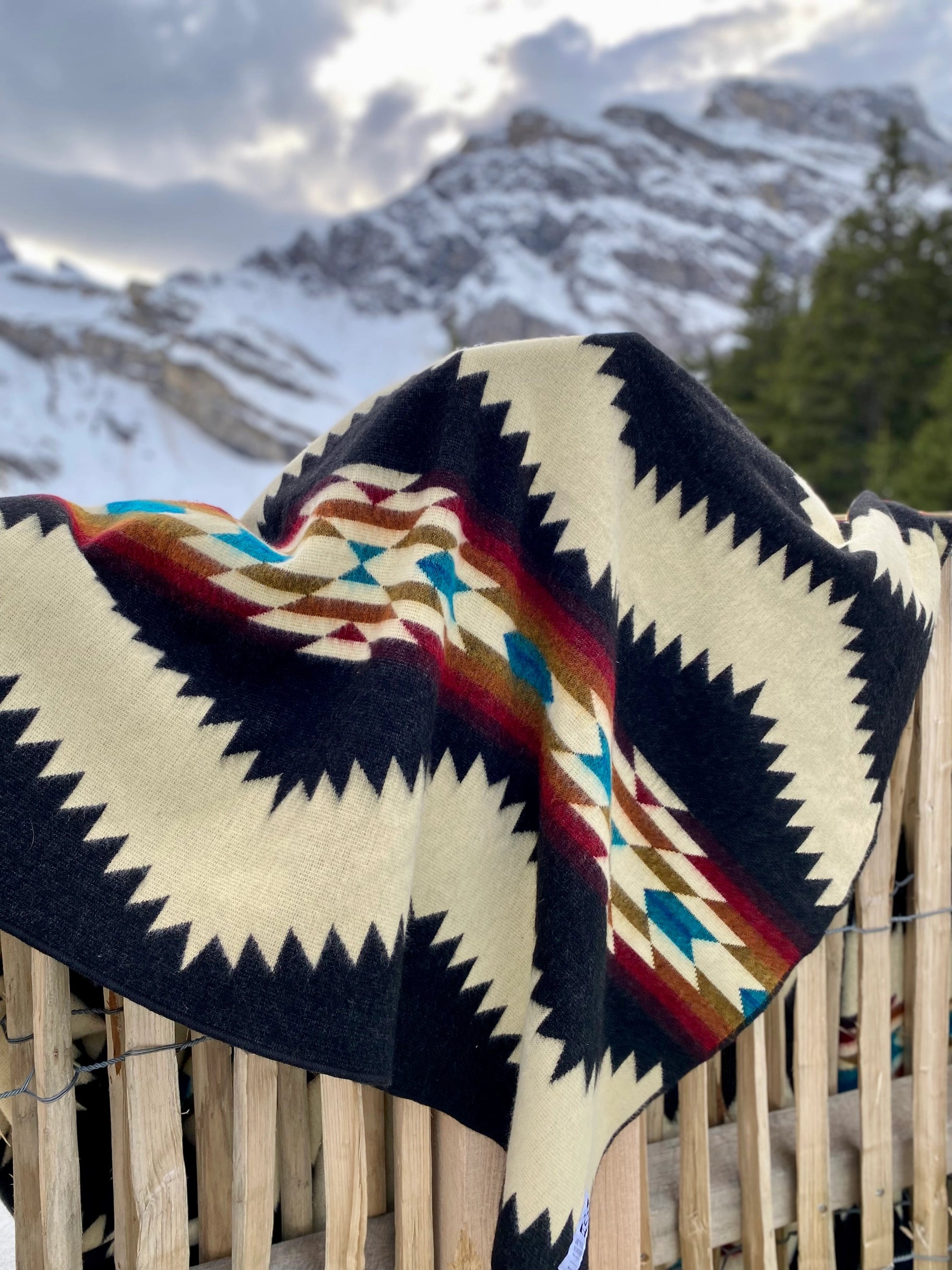 The Tsachila Blanket by Faire, featuring black, cream, red, blue, and brown geometric patterns, is draped over a wooden fence with snowy mountains and pine trees—ideal for boho home decor.