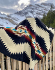 The Tsachila Blanket by Faire, featuring black, cream, red, blue, and brown geometric patterns, is draped over a wooden fence with snowy mountains and pine trees—ideal for boho home decor.