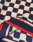 The Checkerboard Silk Bandana Americana by Faire is a folded silk scarf made from 100% silk twill that features a geometric pattern of navy squares and red dots on a beige background, accented with red trim. This elegant accessory includes hand-rolled edges, adding a touch of sophistication to its classic design.