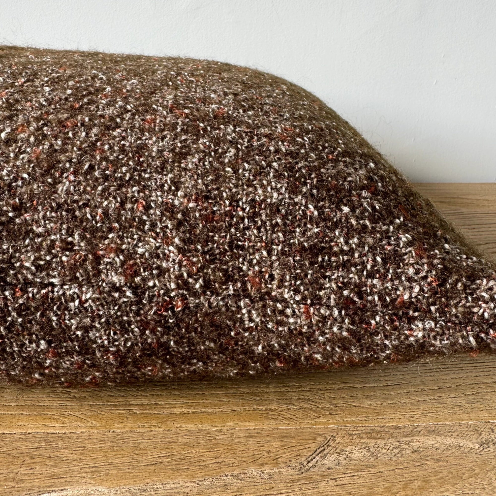 The Adah Coco Wool & Linen Pillow by Faire features a textured coco brown stonewash finish, shown in close-up as it rests on a wooden surface with a plain light-colored wall in the background.