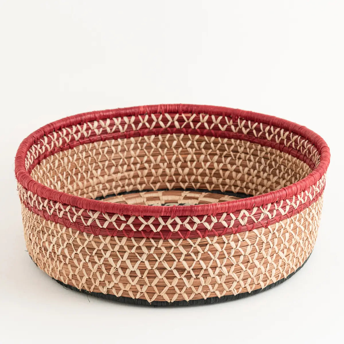 The Large Manuela Basket by Faire is a round, handwoven piece with natural and red crisscrossed fibers, a red-wrapped rim, and a black base. Stylish and decorative, it stands out beautifully against a white background.