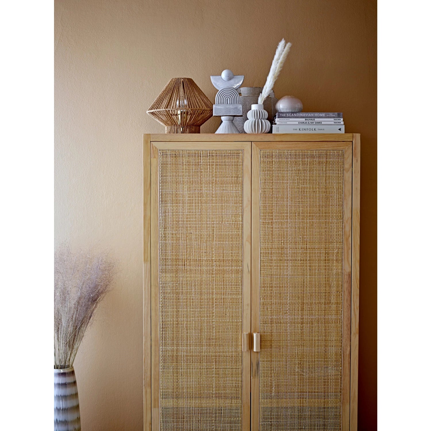 A tall wooden cabinet with rattan doors against a warm beige wall in a Scottsdale, Arizona bungalow, decorated with a stack of books, two small sculptures, and a Bloomingville Pleated Vase atop it. A vase with dried grasses is placed to the left side on the floor.