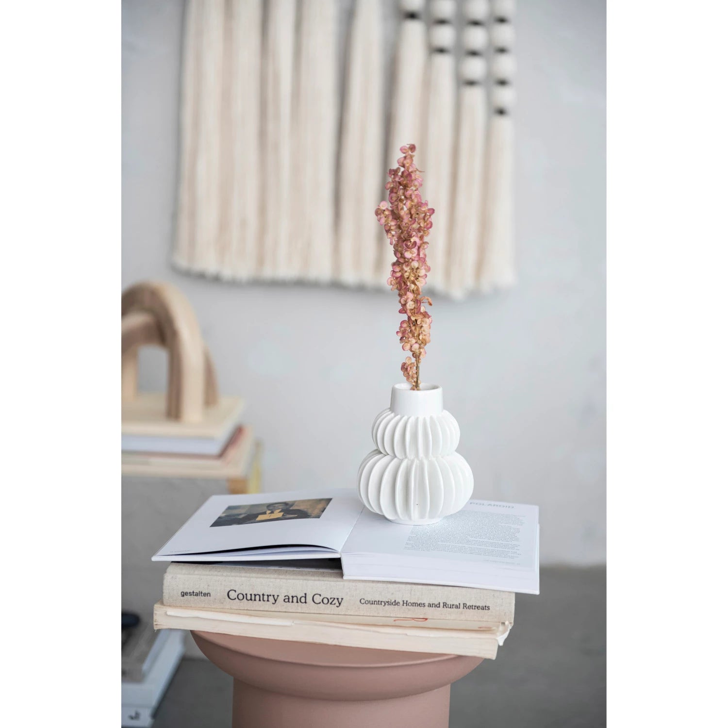 A stylish white Bloomingville Pleated Vase with a textured design holds dried pink flowers, placed on an open book titled "Bungalow and Cozy". The setting includes a neutral-toned background with a woven wall piece.