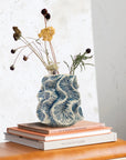 A Textured Vase by Bloomingville, filled with dried flowers, highlights artisanal craftsmanship as it rests on books with white and orange covers against a neutral wall background.