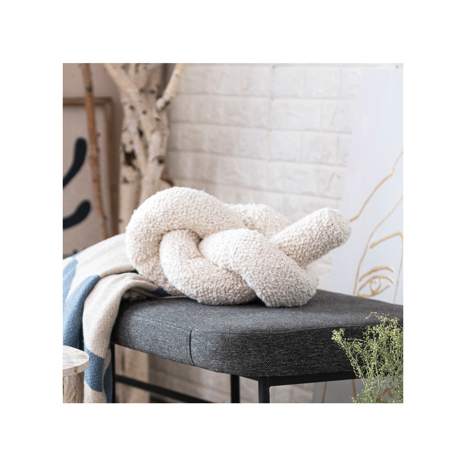 A Bloomingville Cotton Knotted Pillow in cream sits on a dark gray bench beside a blue and white knit blanket, with a white brick wall, drawing, and decorative branches in the background.