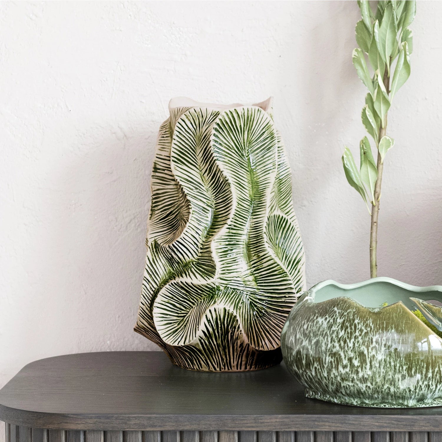 The Bloomingville Green Floral Texture Vase (round stone vase) with a green palm leaf pattern sits on a gray console table beside a green glazed bowl and a tall leafy stem, set against a textured white wall.