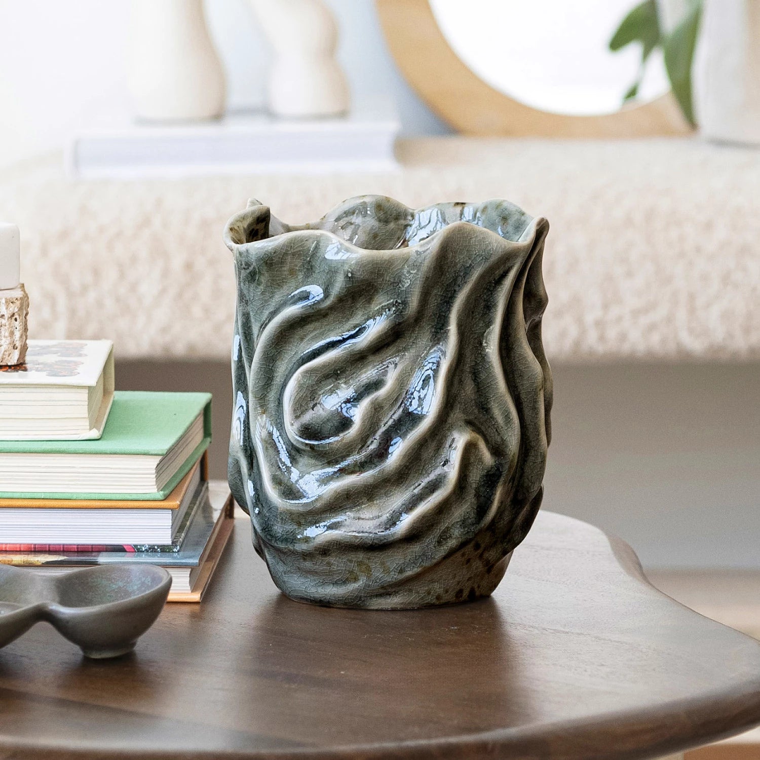 The Bloomingville Wavy Blue Stone Vase (Round Stone Vase), featuring a textured, glossy finish, sits on a wooden table beside stacked books and a small dish in a softly lit, cozy living room.